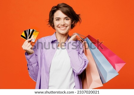 Young happy woman wearing purple shirt casual clothes hold in hand discount cards paper package bags after shopping isolated on plain orange color background studio. Black Friday sale buy day concept