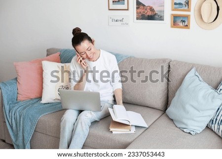 Young woman working on laptop. Millennial girl working remote job from home, using mobile phone, sitting on the cozy sofa, smiling. 
