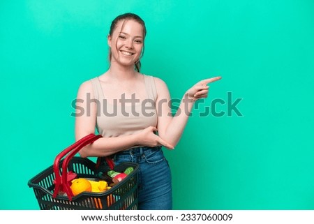 Young woman holding a shopping basket full of food isolated on green background pointing finger to the side