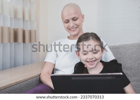 Happy bald woman with alopecia smiles and joyfully spends time with her child watching cartoons on a tablet.