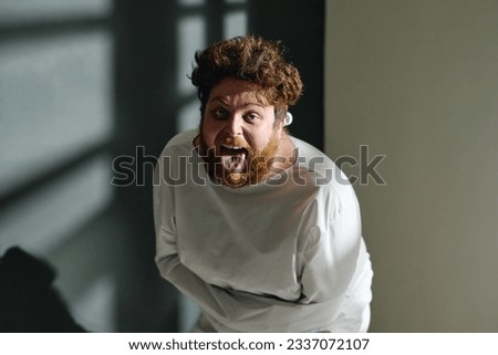 Young man in straitjacket showing tongue and screaming while standing in front of camera in mental hospital ward and looking at you Royalty-Free Stock Photo #2337072107