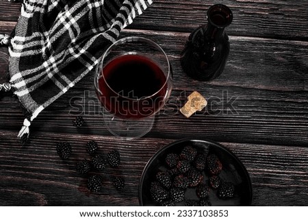 Red wine in a glass next to a black bottle and some blackberries on a wooden table in a dark moody rustic atmosphere, view from above