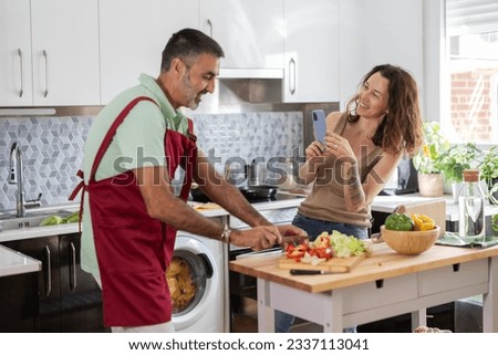 senior couple cooking happy. The woman is recording her partner with the mobile while he prepares lunch