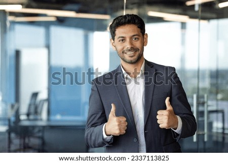 Portrait of young arab businessman, man inside office at workplace standing near window, showing thumbs up very satisfied with financial results of achievement.