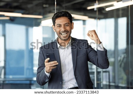 Portrait of successful financier investor, hispanic man smiling and looking at camera, received online win message, man holding hand up gesture of triumph and winner, inside office at workplace. Royalty-Free Stock Photo #2337113843