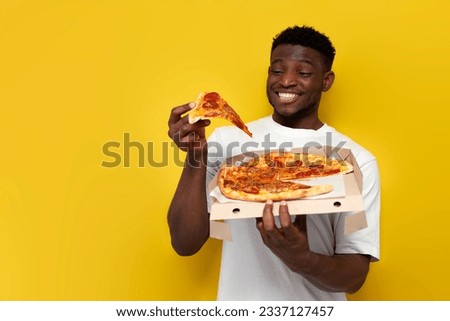 joyful african american man in white t-shirt holds box of pizza and smiles on yellow isolated background, the guy eats fast food and takes slice of pizza Royalty-Free Stock Photo #2337127457