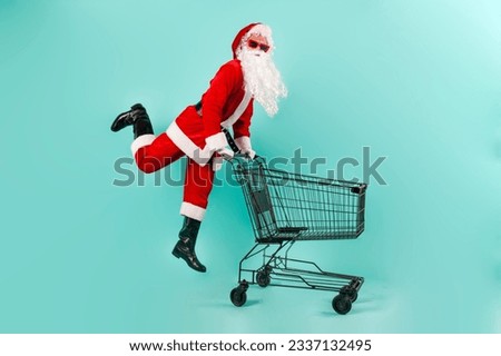 santa claus shopper in sunglasses and suit jumps and flies with empty shopping cart to the store on blue isolated background, santa rushes to the supermarket for shopping for new year and christmas