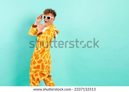 young funny guy in orange giraffe pajamas dances at party in sunglasses and points back on blue isolated background, man in animal costume shows on the copy space from behind, pajama disco concept