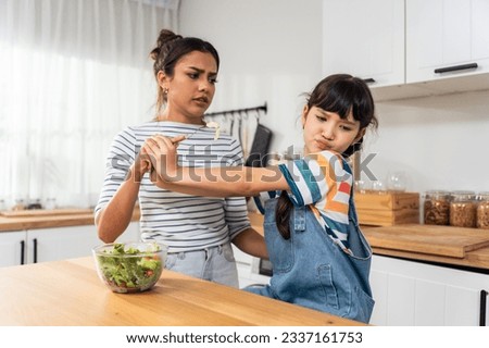 Caucasian mother teach and motivate young daughter eat green vegetable. Adorable child shake head say no to mom, don't want to eat salad on dinner plate in kitchen. Healthy food and childhood concept.