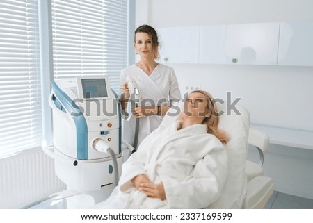 Portrait of female beautician specialist standing posing with device for photo rejuvenation in cosmetology clinic, looking at camera. Woman client receiving stimulating electric facial treatment.