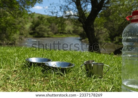 Close-up of aluminum plates and mugs stand on the green grass against the incredible landscape of a mountain river and trees on a salty day. Cooking while traveling by car.