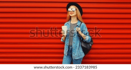 Portrait of young woman with cup of coffee wearing black round hat, denim jacket and backpack on red background