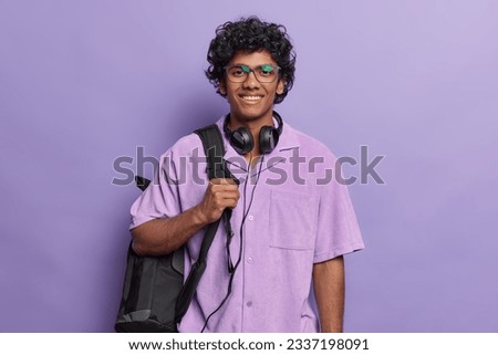 Cheerful millennial Hindu student poses over purple background dressed casually smiles pleasantly being on way to university with rucksack uses headphones for audio lessons. Studying and education