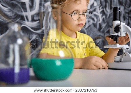 Focused little boy conducts an experiment in real laboratory against the background of chalkboard. In a chemistry lesson, child who is interested in science studies molecules using a real microscope.