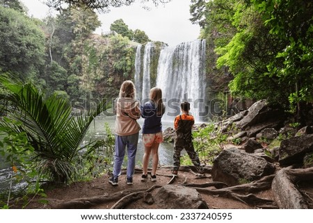Three people, a mum and two children at an idyllic location looking at a waterfall, with their backs turned. This was taken at Whangarei Falls, New Zealand. Royalty-Free Stock Photo #2337240595