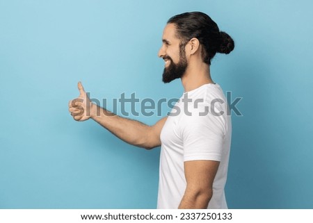 Side view of man with beard wearing white T-shirt standing with thumbs up, like gesture, demonstrating approval and agree with suggestion. Indoor studio shot isolated on blue background. Royalty-Free Stock Photo #2337250133