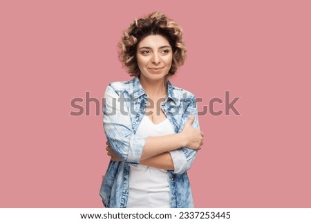 Portrait of positive pretty young adult woman with curly hairstyle wearing blue shirt standing with folded hands, looking away and dreaming. Indoor studio shot isolated on pink background. Royalty-Free Stock Photo #2337253445