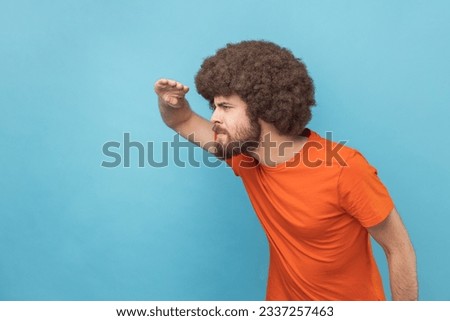 Side view of man with Afro hairstyle wearing orange T-shirt looking far away at distance with hand over head, attentively searching for bright future. Indoor studio shot isolated on blue background. Royalty-Free Stock Photo #2337257463