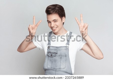 Portrait of young brunette man standing makes peace gesture, shows v sign, looking at camera with toothy smile, wearing denim overalls. Indoor studio shot isolated on gray background.