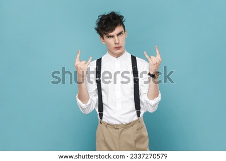 Portrait of serious young attractive man wearing white shirt and suspender, standing looking at camera and showing rock and roll gesture. Indoor studio shot isolated on blue background.