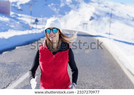 Smiling young female blonde in red vest beanie cap polarized sunglasses looking at camera, while jogging on asphalt road near snow against blurred mountain with clouds