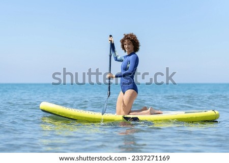 Side view of female surfer in swimsuit kneeling on paddleboard on blue sea while paddling with both hands and looking at camera in bright daylight
