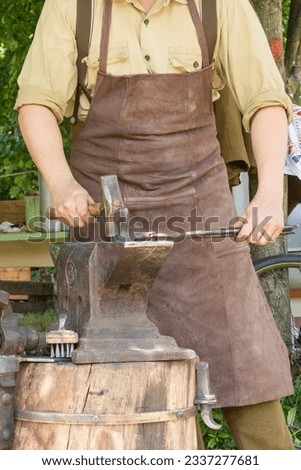A blacksmith in a brown leather apron is forging a piece of iron on an anvil