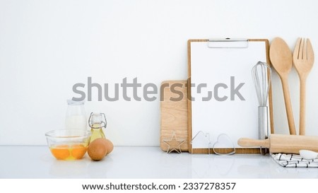 Close-up picture of fresh baking ingredients, eggs, milk, and oil and kitchen wares, utensils on white background table and wall with recipe notebook for making delicious pastry.
