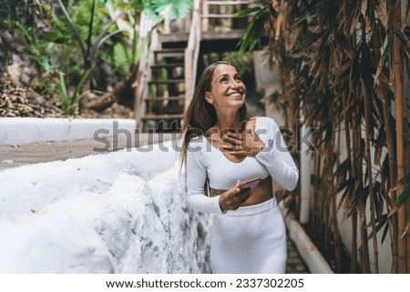 Smiling young female freelancer with long hair in white clothes walking along narrow path near wall with bamboo while surfing internet on smartphone and working remotely on project Royalty-Free Stock Photo #2337302205