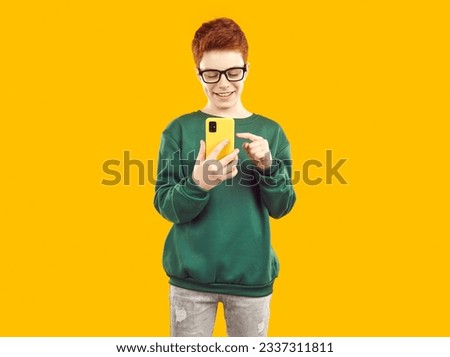 Handsome happy smiling boy with glasses and red hair, dressed in green sweater and gray jeans, flips through the menu on his mobile phone or takes a photo.Studio photo on an isolated yellow background