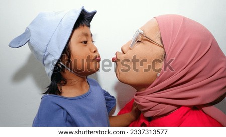 Image of an Asian woman wearing a hijab and her daughter facing each other, the mother posing to kiss her daughter with a happy expression. concept photo of happy mother and daughter. white background