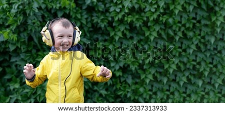 A positive little boy of 6 years old in a yellow jacket and yellow retro earphones listens to music. The boy stands against the background of a wall of plants. Banner with space for text