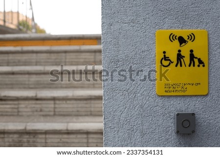 Yellow sign with the sign of a person with a disability in a wheelchair, a person with baby stroller and braille text next to a gray button for calling for help on the stairs in front of the entrance