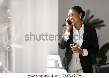 Happy black woman in suit having phone conversation and drinking coffee to go, female boss standing in office in the morning, looking at copy space and smiling Royalty-Free Stock Photo #2337369079
