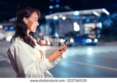 Smiling young woman walking in the city at night with earphones, listening online music from smartphone, illuminated city night lights, side view, free space