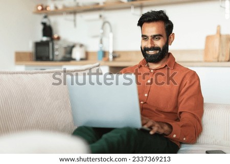 Freelance career and modern technologies. Happy Arab bearded man surfing web on computer at home. Freelancer guy using laptop browsing internet for work purpose sitting on sofa indoor
