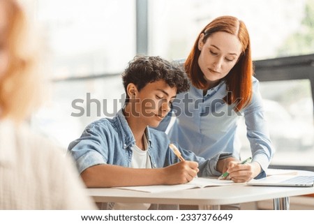 Young pensive school teacher explaining something to smart teenager, standing and showing in notebook, holding pen. Serious tutor assisting helping with homework, exam preparation. Education concept Royalty-Free Stock Photo #2337376649