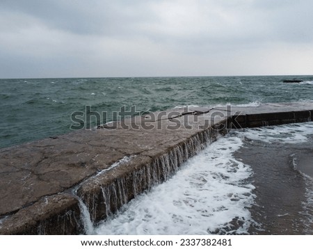 The wave rolls over a large square rock lying on the shore, covered with green seaweed. In the cloudy weather the summer. The foam on the wave from the slab over the pier. High quality photo