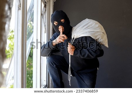 Thief in black clothes wearing a mask carrying a large bag of loot over his shoulder trying to escape from window and looking at camera making hush and mute gesture 
