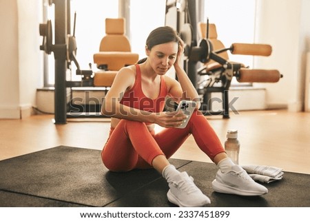 Fit female athlete in sportswear sitting on mat and using fitness app between workout sets in gym Royalty-Free Stock Photo #2337451899
