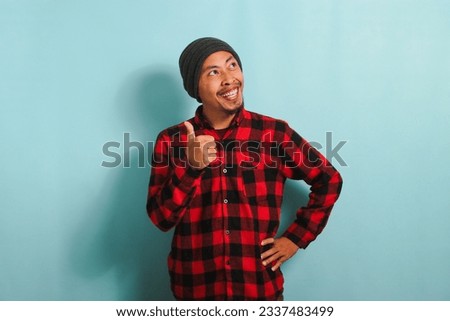 A confident young Asian man with a beanie hat and a red plaid flannel shirt is giving a thumbs-up while looking at right copy space, expressing a positive review. He is isolated on a blue background