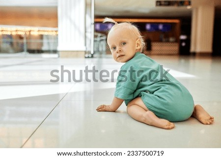 A small child crawling on the floor in a public place, shopping mall. Tiny cute blonde girl in green clothes having fun with mom while shopping. Funny weekend, happy childhood.
