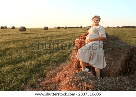 blond pregnant woman in white dress with basket on hay summer field background