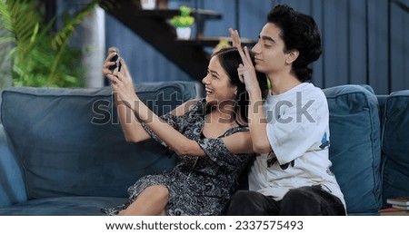 Smiling girlfriend sitting on sofa taking funny faces selfie with teen boy using mobile phone at indoor home. Happy Indian teenager girl making self photos record vlog on smartphone enjoy fun together Royalty-Free Stock Photo #2337575493