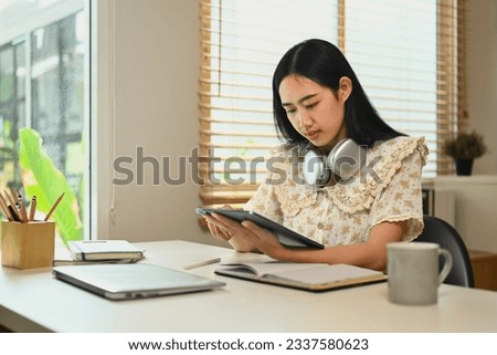 Pretty young Asian woman using digital tablet and making notes, browsing internet, remote work and stying online