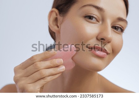 Crop photo of a young adult woman doing face self-massage with pink jade for spa skin care treatment. Beauty face skin care. Girl using natural massager tool on blue background