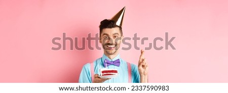 Holidays and celebration concept. Happy young man enjoying birthday party, wearing cone hat and cross fingers, making wish on bday cake with candle, pink background.