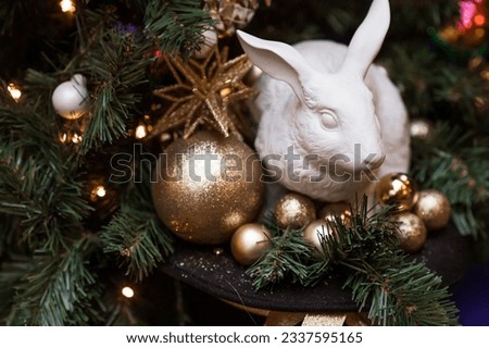 Beautiful festive decorations on the Christmas tree in golden tones and a white rabbit in a hat. Beautiful Christmas background. New Year's card 2024.