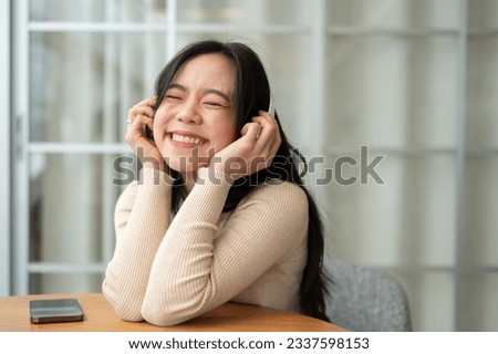 A portrait of a beautiful and happy young Asian woman is sitting at a table indoors and enjoying the music on her headphones. Hobby and lifestyle concept