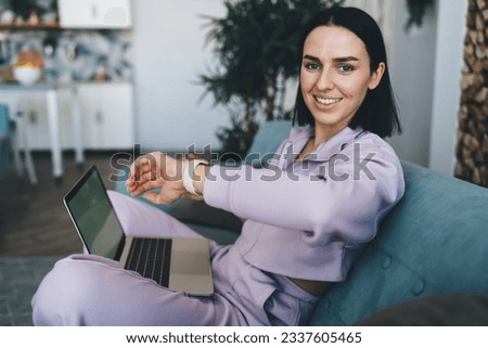 Positive young female showing smartwatch with raising hand and smiling while looking at camera and relaxing on sofa with laptop in blurred living room at home